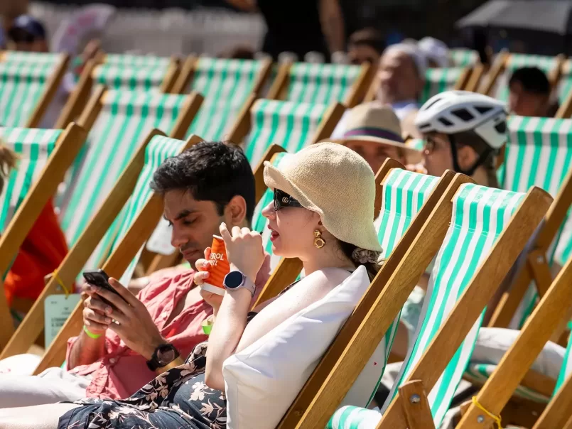 People relax on striped deck chairs outdoors on a sunny day. A woman wearing a sunhat and sunglasses drinks from a bottle, while a man beside her looks at his phone. Other people lounge and chat in the background.