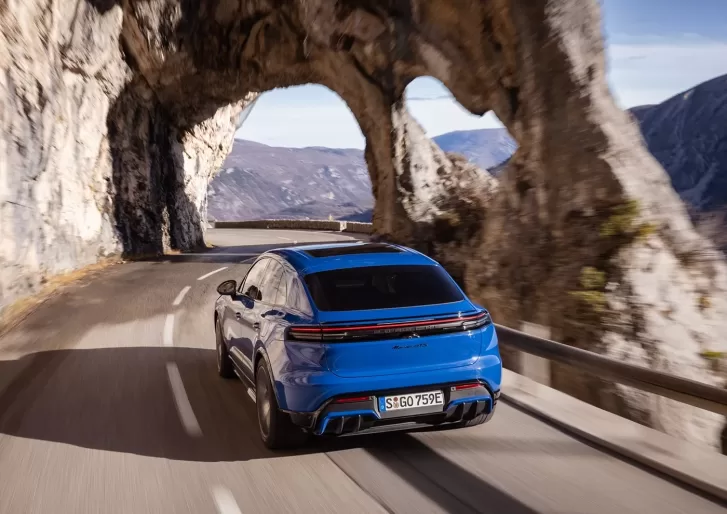 A blue sports car drives along a winding mountain road, passing through a natural rock arch with scenic cliffs and distant hills in the background under a partly cloudy sky.