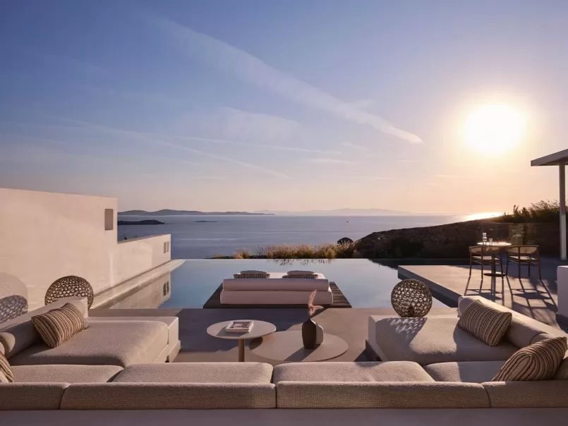 Modern outdoor lounge area with plush seating and a coffee table beside an infinity pool, overlooking the ocean and distant hills at sunset. Warm sunlight bathes the scene, creating a tranquil and luxurious atmosphere.