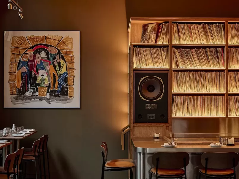 A cozy restaurant interior with a large painting on the wall, shelves filled with vinyl records, a prominent speaker, and neatly set tables and chairs under warm lighting.