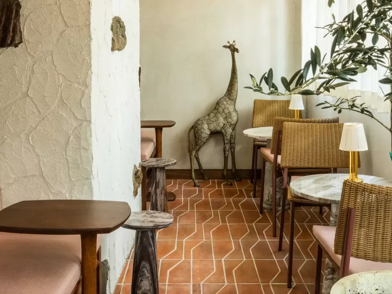 A cozy cafe corner with tiled floors, wooden tables, wicker chairs, potted plants, and a decorative giraffe statue against a cream-colored wall.