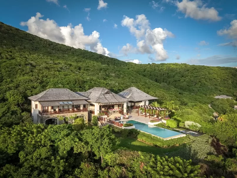 A large luxury villa with a pool sits surrounded by lush green hills under a blue sky with scattered clouds. The house features multiple roofs and balconies, with landscaped gardens and outdoor seating areas.