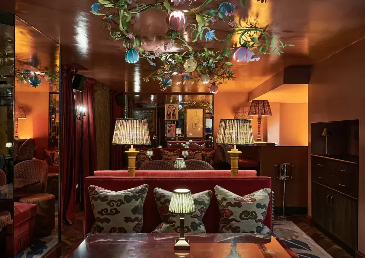 A cozy, dimly lit lounge with red velvet sofas, patterned pillows, and table lamps. Floral light fixtures hang from the ceiling, and mirrors and warm amber lighting create a rich, inviting atmosphere.