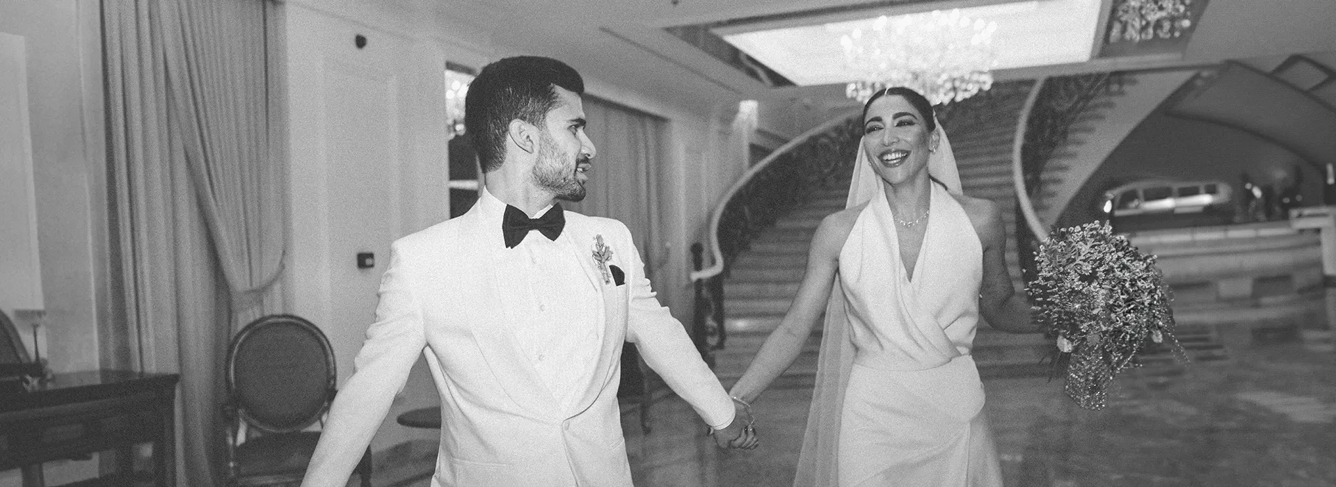 Black and white photo of smiling groom and bride wearing wedding dress  in a grand room.