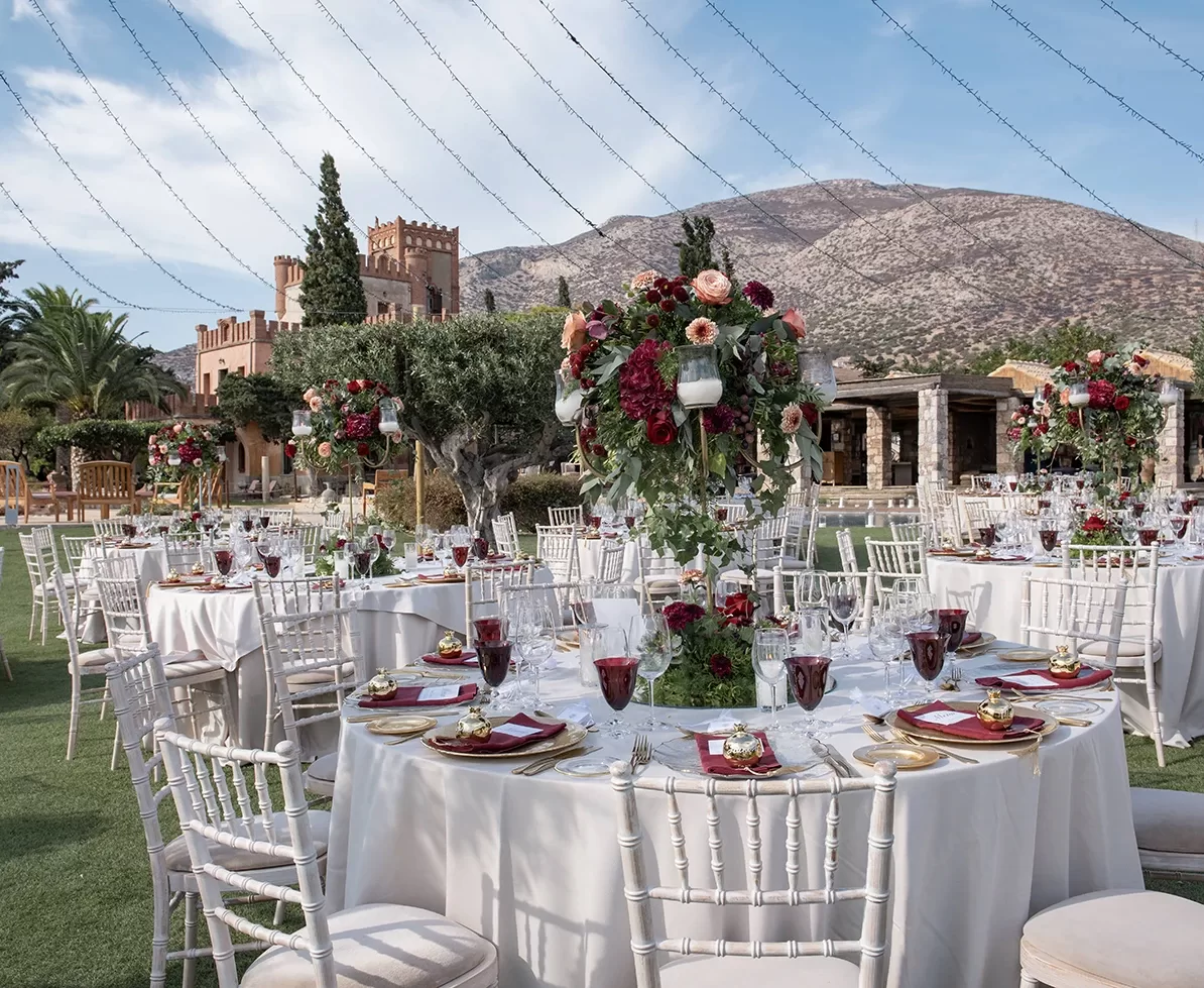 Outdoor wedding reception with round tables adorned with elegant centerpieces of red and pink flowers. White chiavari chairs surround the tables. A castle-like building and mountains are visible in the background under a clear sky.