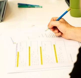 A person is writing on a worksheet with several highlighted sections on a desk. A pen and notebook are visible in the background.