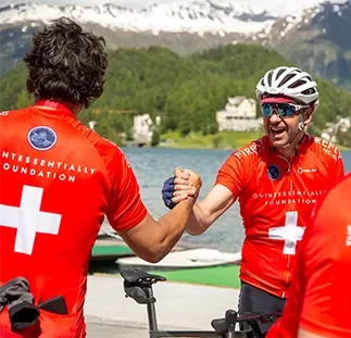 Two cyclists in red jerseys with white crosses shake hands by a scenic lake with snow-capped mountains in the background. They appear cheerful and are part of a group. The jerseys have foundation logos on them at Quintessentially Foundation's annual bike ride challenge.
