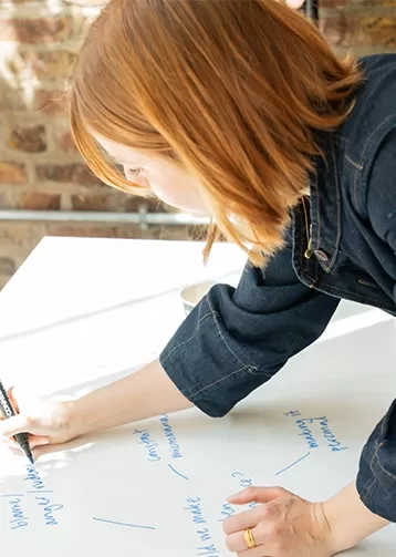 A staff member of Quintessentially Foundation with shoulder-length red hair writes on a large sheet of paper using a blue marker. Theyre wearing a dark denim shirt and focusing on their work. The background shows a brick wall and natural lighting.