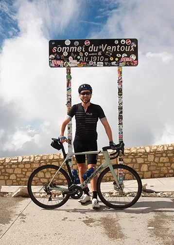 A cyclist in black gear stands with a road bike at the summit of Mont Ventoux. Behind them is a sign reading Sommet du Ventoux Alt. 1910 m, covered in stickers. The background shows a partly cloudy sky at Quintessentially Foundation's annual bike ride challenge.