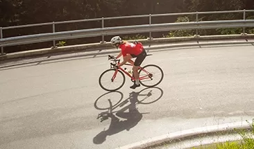 A cyclist in a red shirt and helmet rides a road bike on a sunlit, curved roadway bordered by a metal guardrail, surrounded by trees. The sun casts a clear shadow of the cyclist on the pavement at Quintessentially Foundation's annual bike ride challenge.