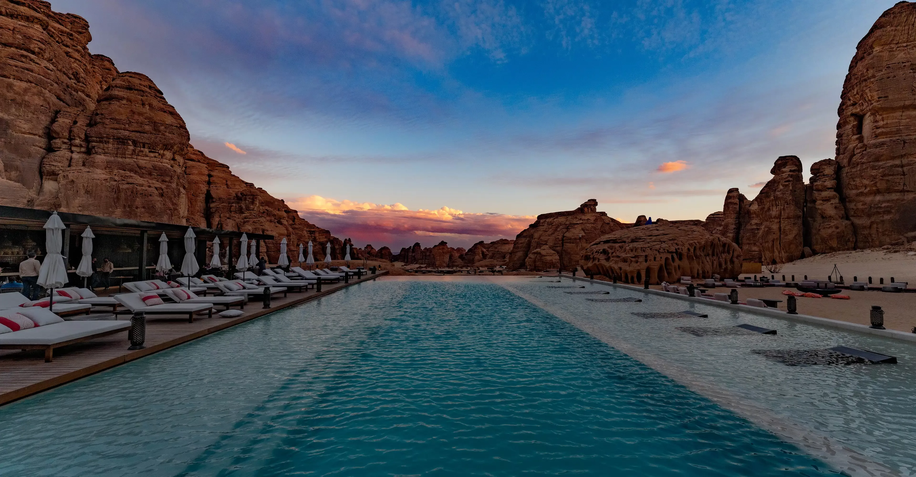 A tranquil pool stretches between rocky desert cliffs at sunset, lined with lounge chairs and umbrellas on both sides, with a dramatic sky overhead.