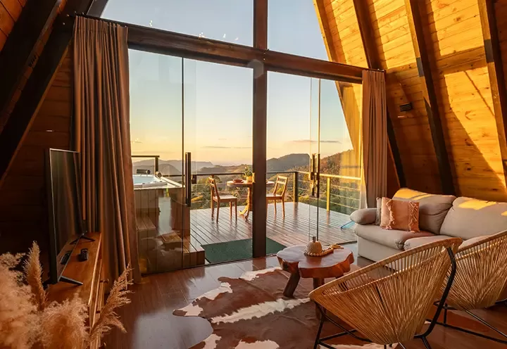 Cozy wooden cabin interior with large glass doors opening to a deck, hot tub, and scenic mountain view at sunset. Room features a sofa, cowhide rug, chairs, TV, and decorative plants. Warm natural light fills the space.