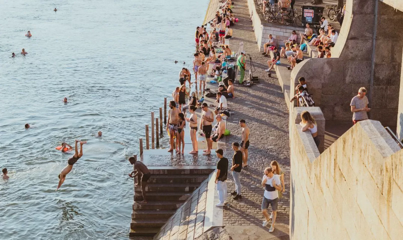 People gather along stone steps and a walkway beside a river; some are swimming, diving, or standing in the water, while others sit or relax on the steps in the sun.