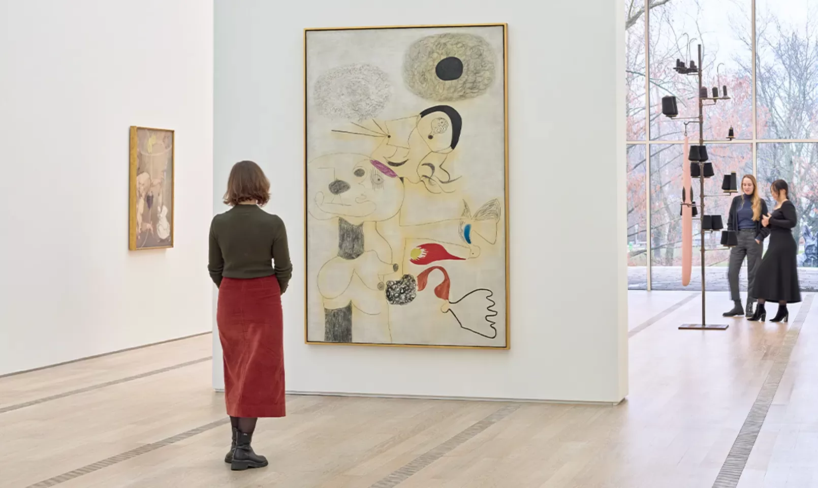 A person stands in a gallery viewing a large abstract painting. Two other people talk near a modern sculpture by large windows, and another painting hangs on a nearby wall. Light streams in from outside.