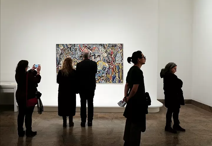 Several people stand in a gallery observing an abstract painting on a white wall; one person photographs the artwork, while others look at it or walk by in different directions.