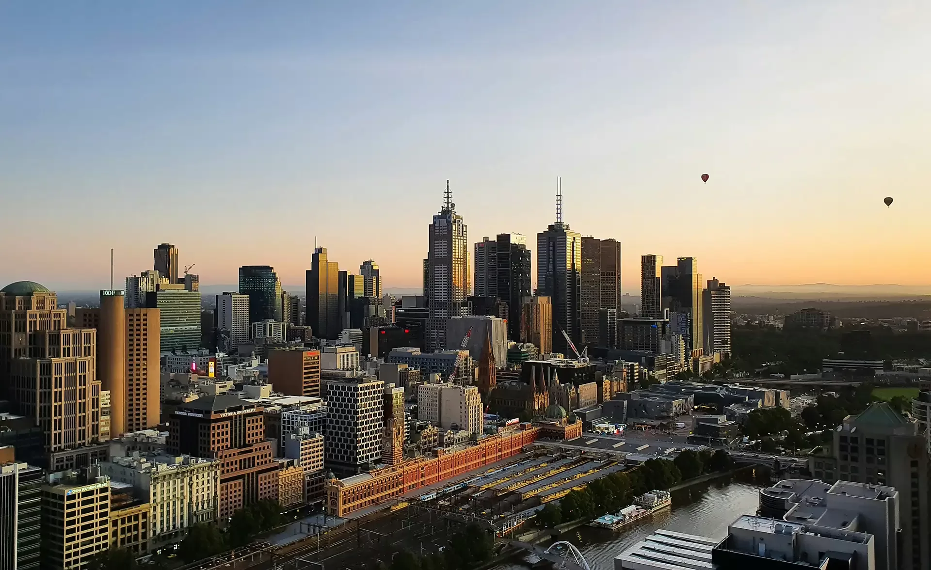 The Melbourne skyline at sunset, featuring a picturesque blend of colours and silhouettes of the city's architecture and floating hot air balloons.