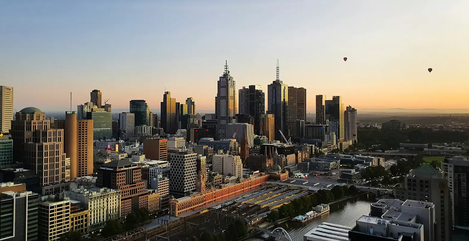 The Melbourne skyline at sunset, featuring a picturesque blend of colours and silhouettes of the city's architecture and floating hot air balloons.