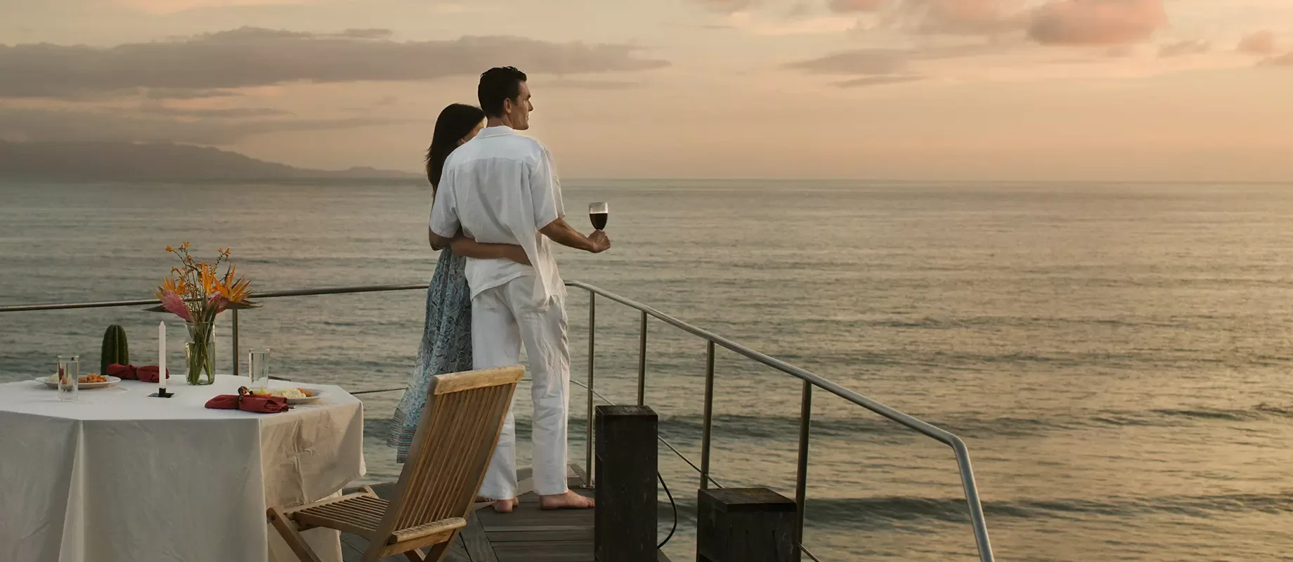 A couple stands on a deck overlooking the ocean at sunset, embracing as they gaze at the view. A table set for dinner and a wooden chair are nearby, creating a romantic atmosphere.
