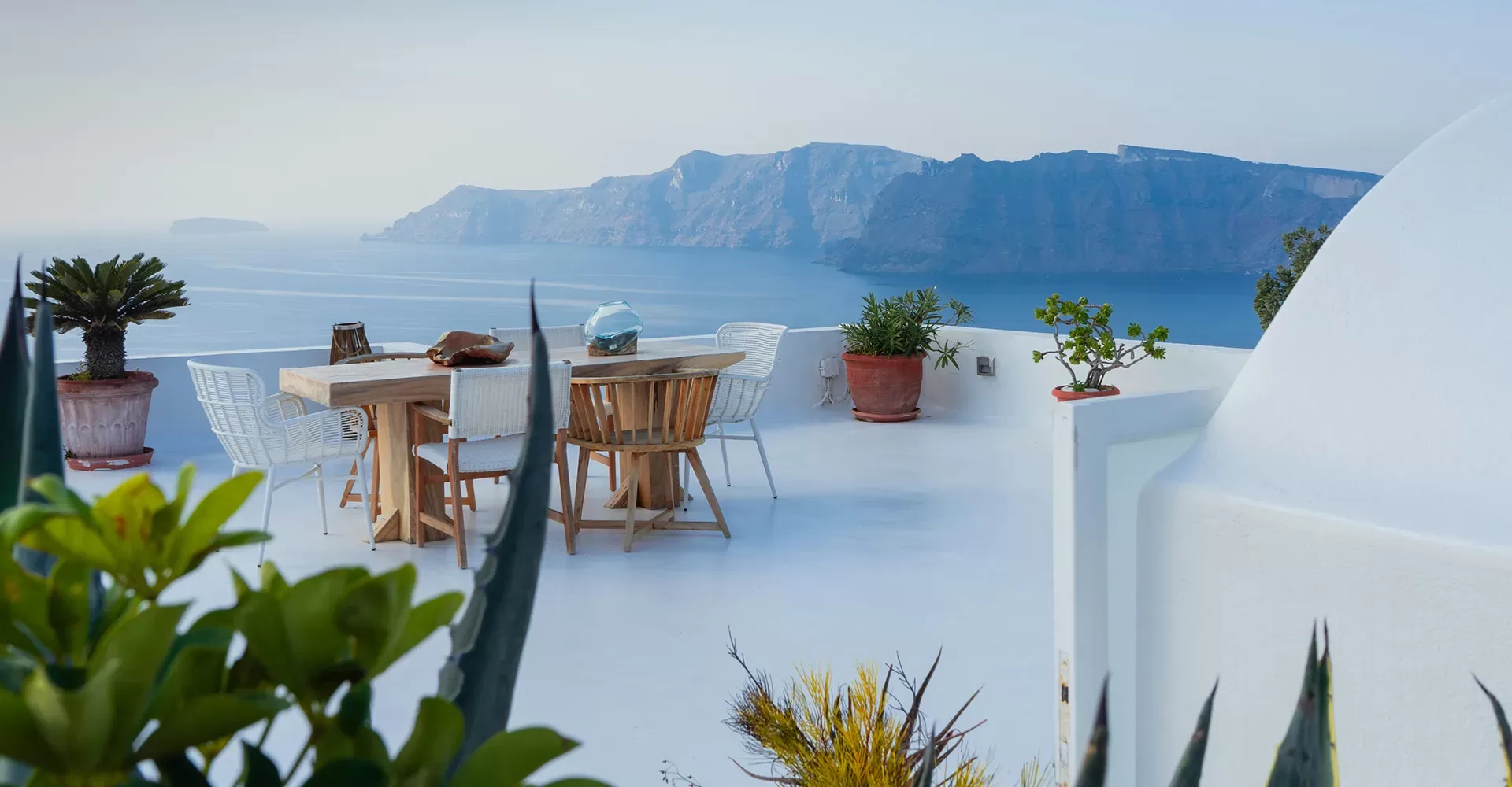A terrace with a wooden dining table and chairs overlooks the calm blue sea, with potted plants and distant mountains under a clear sky, creating a serene, coastal atmosphere.