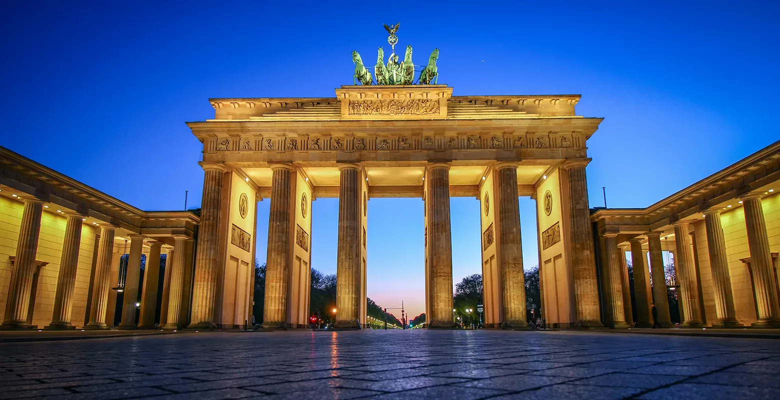 The Brandenburg Gate in Berlin, Germany, illuminated at dusk. The neoclassical monument features six columns and a chariot sculpture on top, with a clear evening sky in the background.