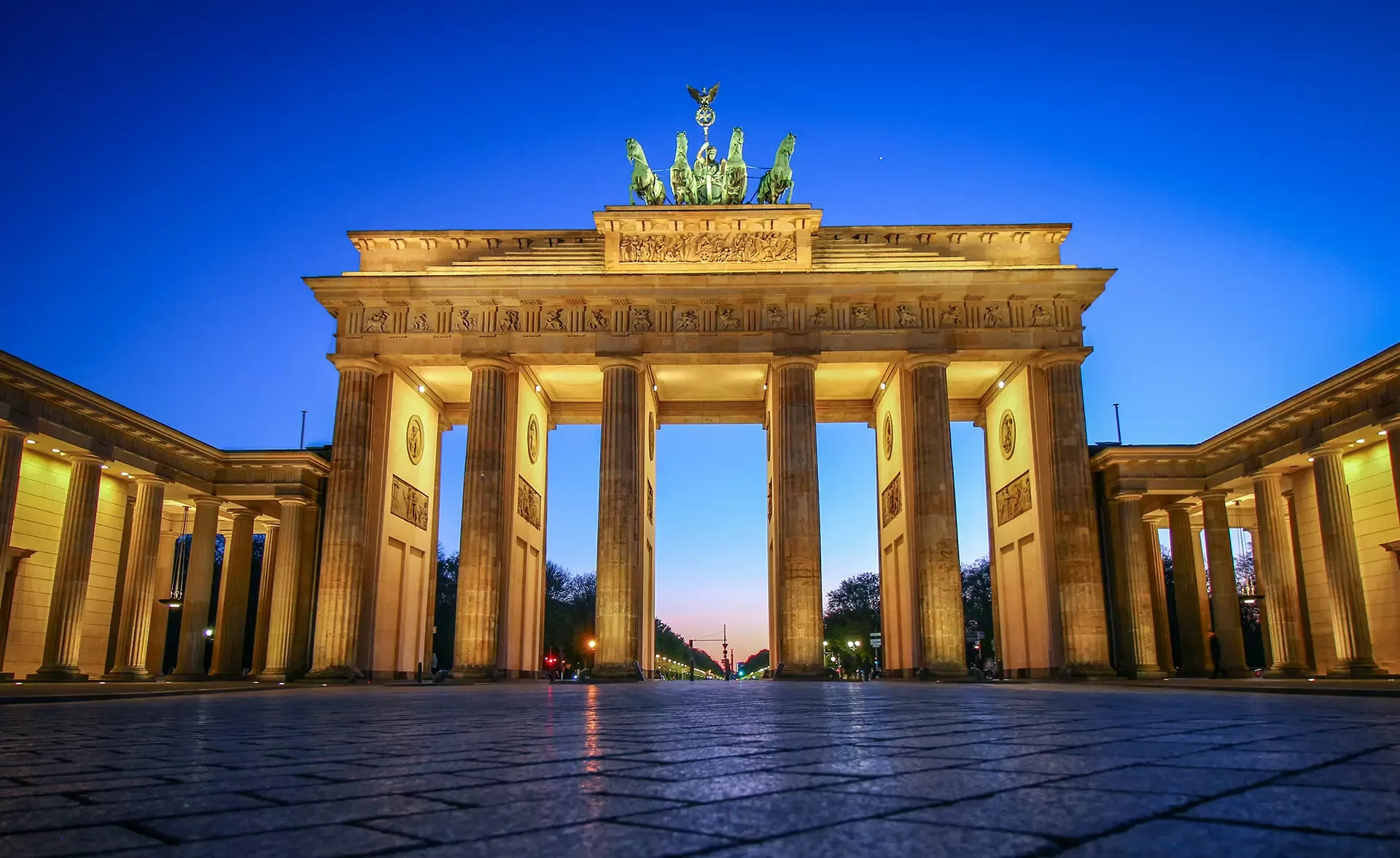 The Brandenburg Gate in Berlin, Germany, illuminated at dusk. The neoclassical monument features six columns and a chariot sculpture on top, with a clear evening sky in the background.