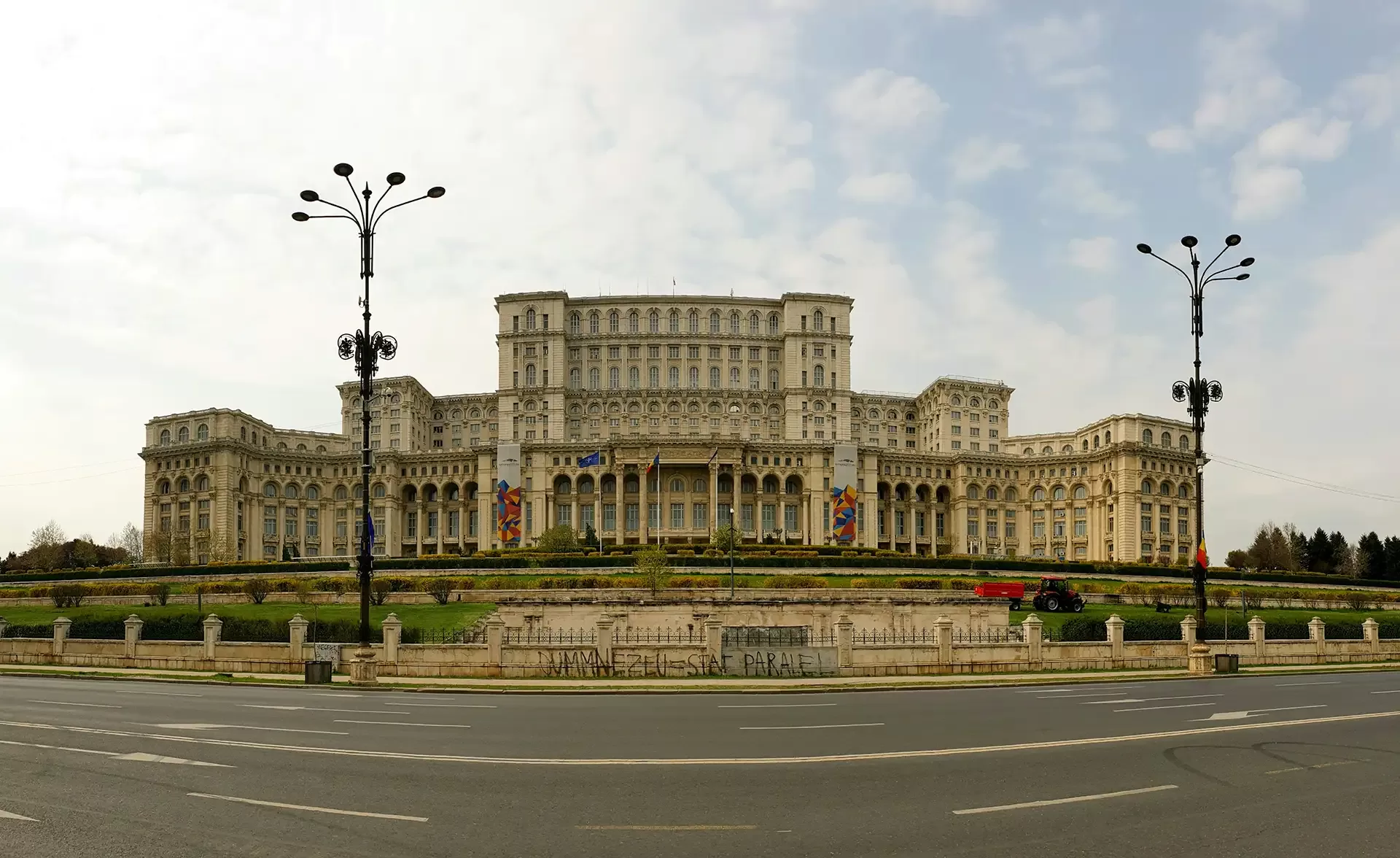 Panoramic view of the massive Palace of the Parliament in Bucharest, Romania, under a partly cloudy sky. The grand neoclassical building is surrounded by manicured lawns and flanked by lamp posts.