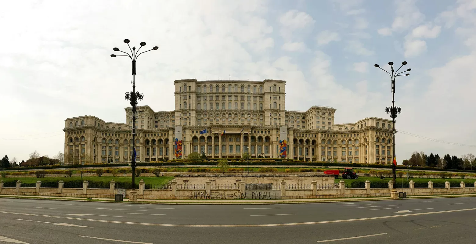 A grand building with a neoclassical design, featuring multiple stories, numerous columns, and a symmetrical facade. It is set against a partly cloudy sky, surrounded by manicured lawns and symmetrical streetlights in the foreground.