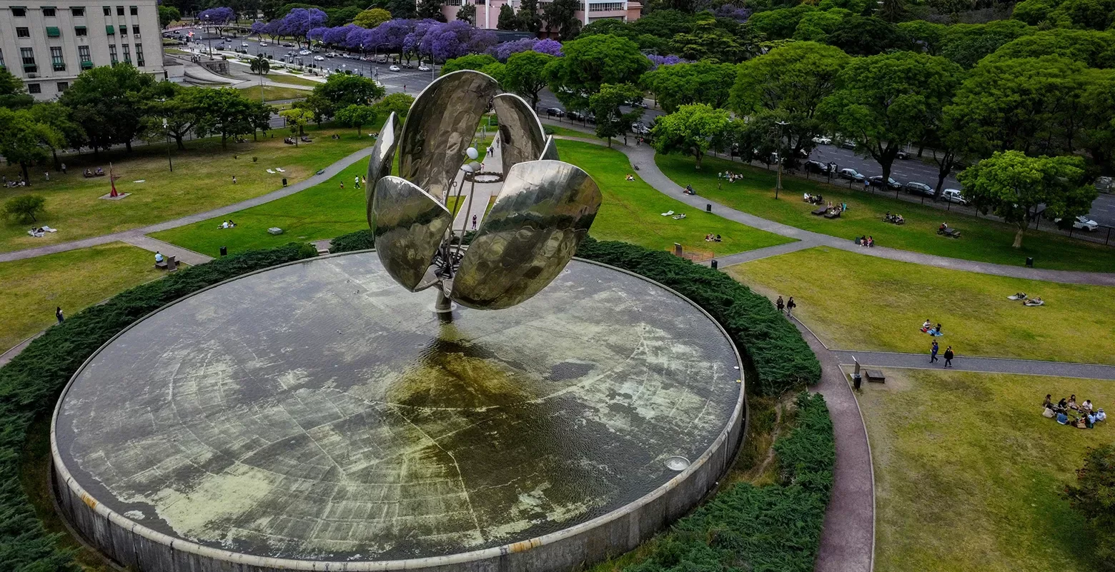 Aerial view of a large metal sculpture shaped like a flower in a circular pond, surrounded by a green park with people sitting on the grass. Trees line the background, and a road borders the park.