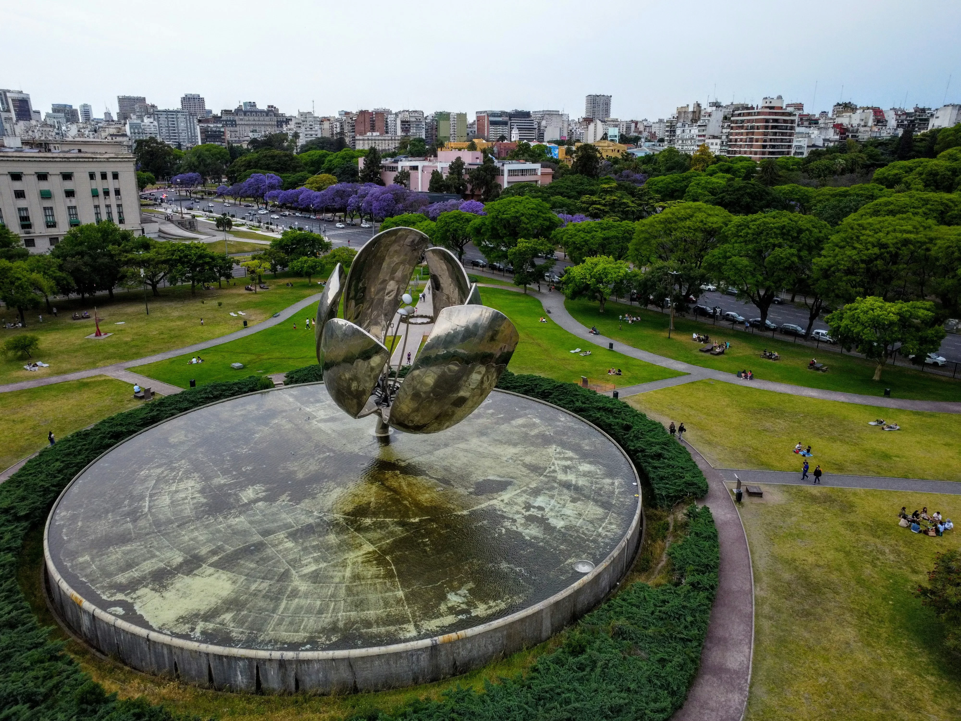 Aerial view of a large metallic flower sculpture in a park, surrounded by green lawns and trees. People are sitting on the grass, and a city skyline is visible in the background.