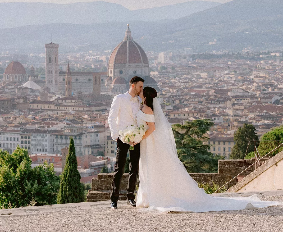 A bride and groom share a kiss on a terrace overlooking Florence, Italy. The bride is in a white gown holding a bouquet, while the groom wears a white suit. The citys iconic dome and historic architecture are visible in the background.