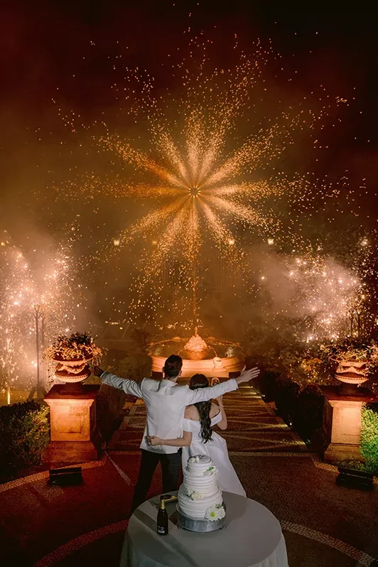 A couple in wedding attire stands with arms raised, facing a vibrant display of fireworks illuminating the night sky. A multi-tiered wedding cake is on the table in front of them. The setting is festive and celebratory.