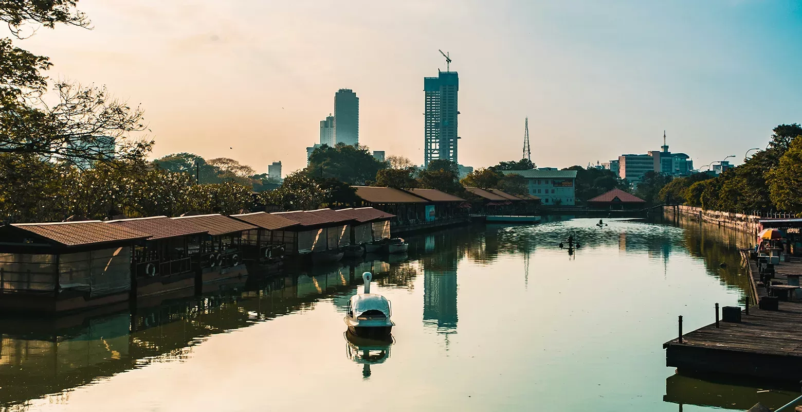 A serene canal at sunset with a swan-shaped pedal boat in the foreground. Lush trees line the waterway, while modern high-rise buildings dominate the background skyline. The sky is a gradient of blues and oranges.