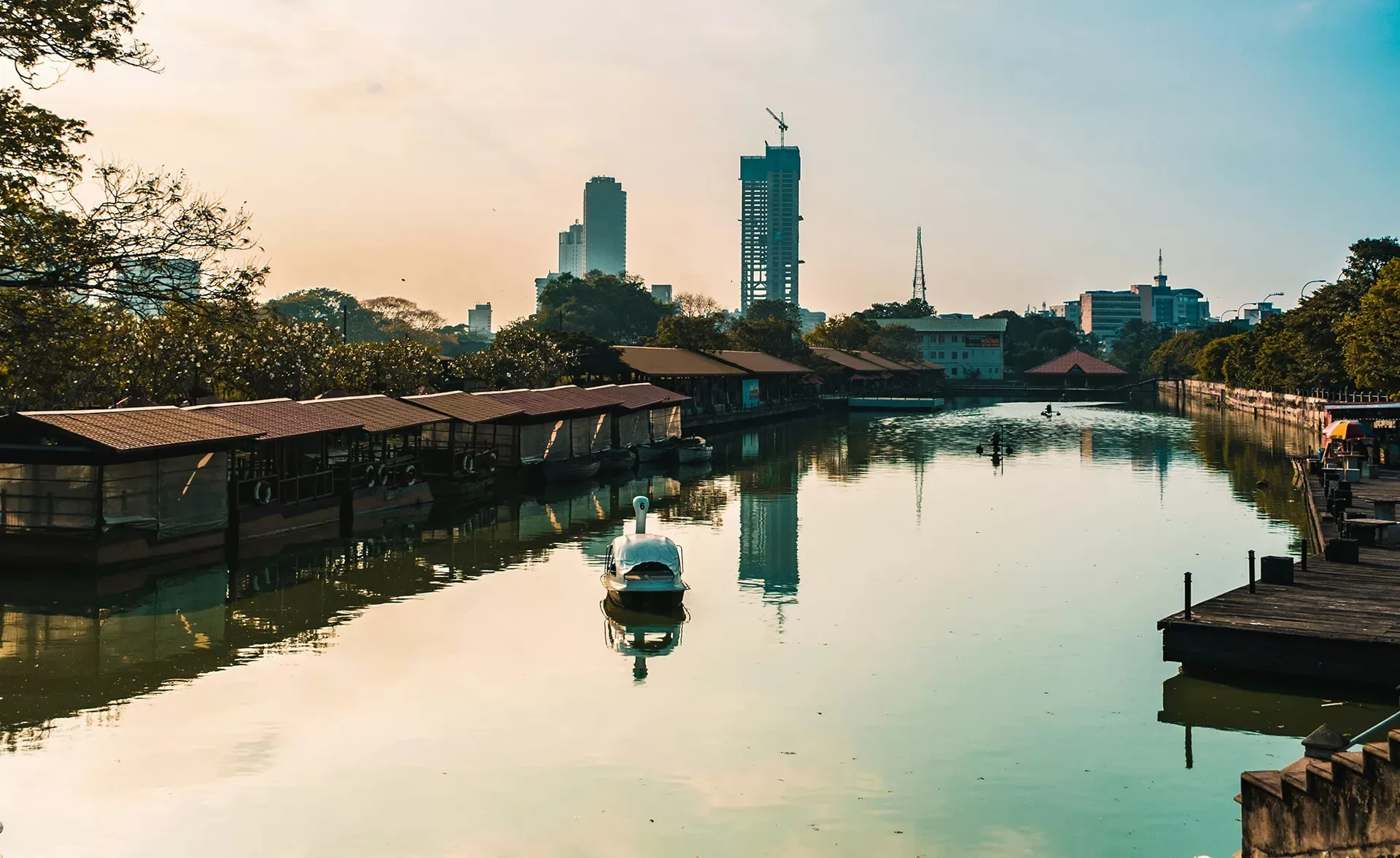 A serene canal scene at sunset with a white boat moored on still water, bordered by trees and wooden structures on each side. In the background, tall city buildings are silhouetted against the orange sky.