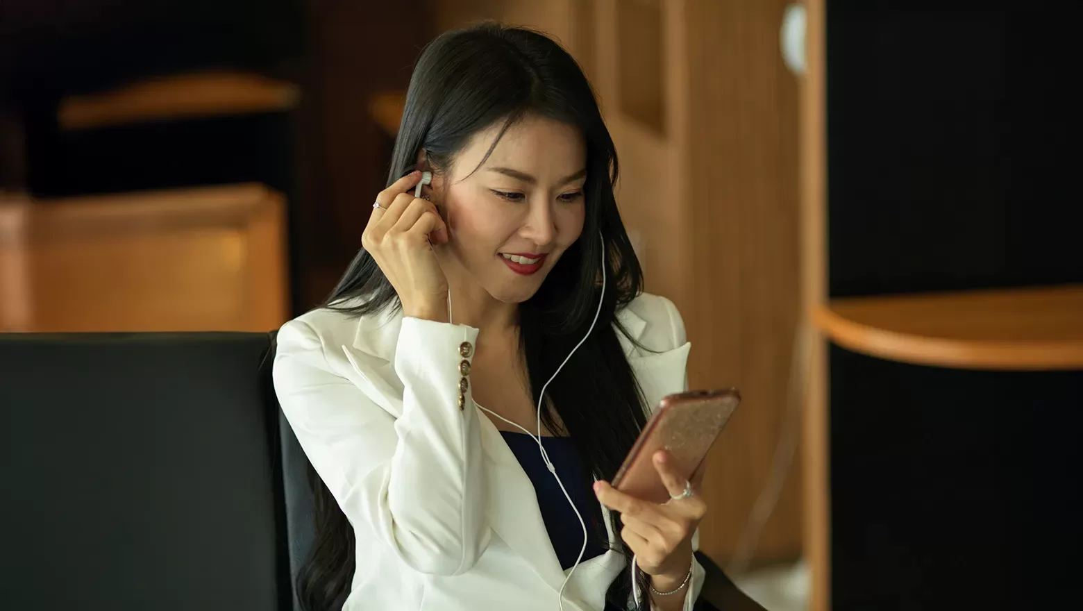 A woman in a white blazer sits indoors, smiling while looking at her smartphone and putting in earphones.
