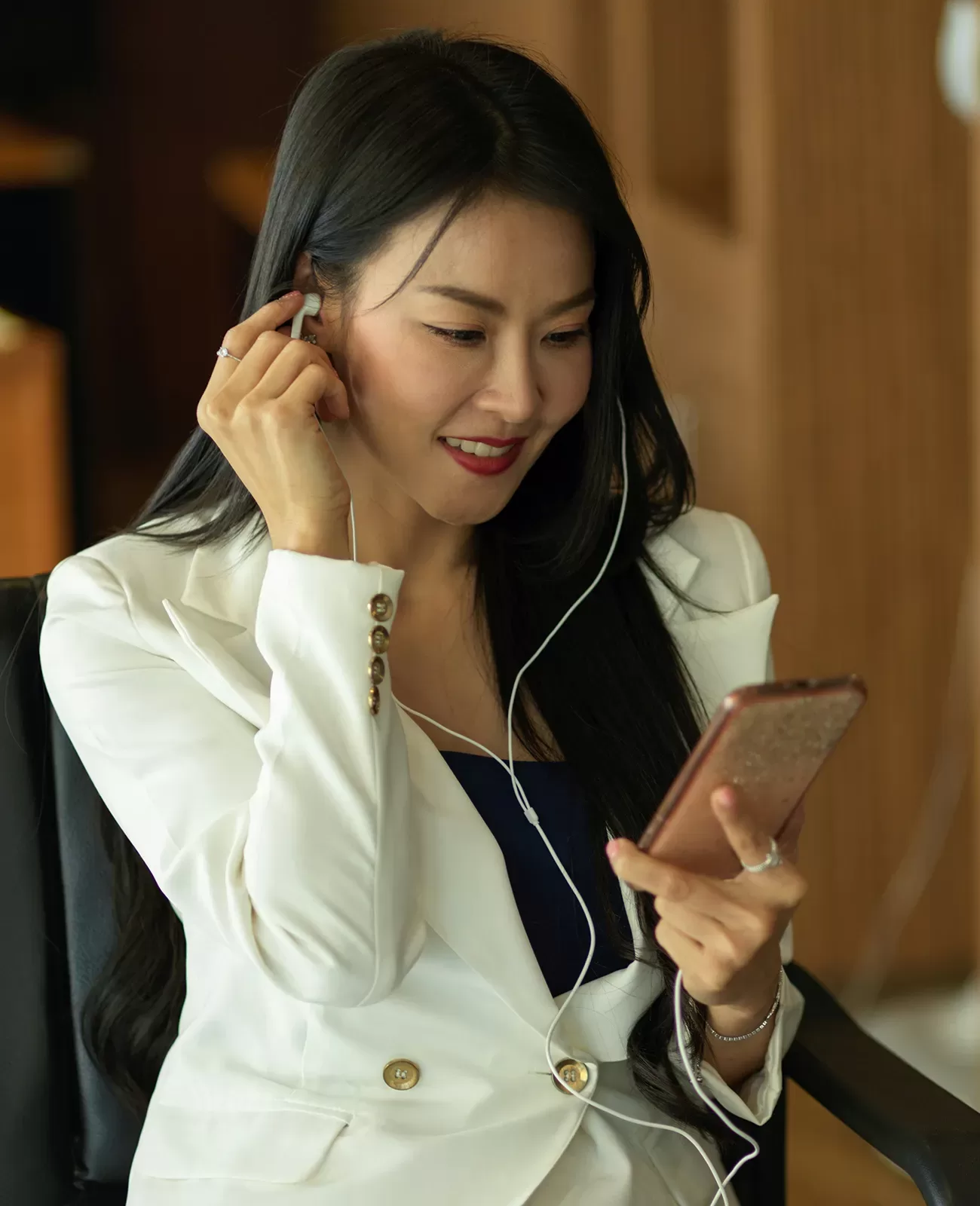 A woman with long black hair wearing a white blazer sits and smiles while looking at her smartphone, listening to something with wired earphones.