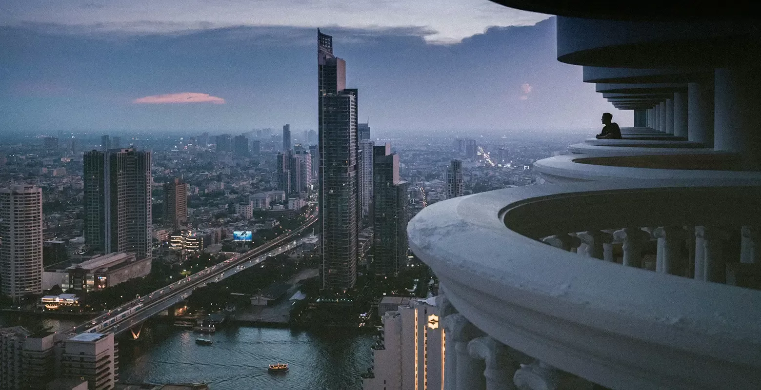 A city skyline at dusk with tall buildings and a river. A person stands on a curved balcony in the foreground, gazing at the urban landscape. The sky is a gradient of blue and purple, enhancing the citys evening lights  in Bangkok, Thailand.