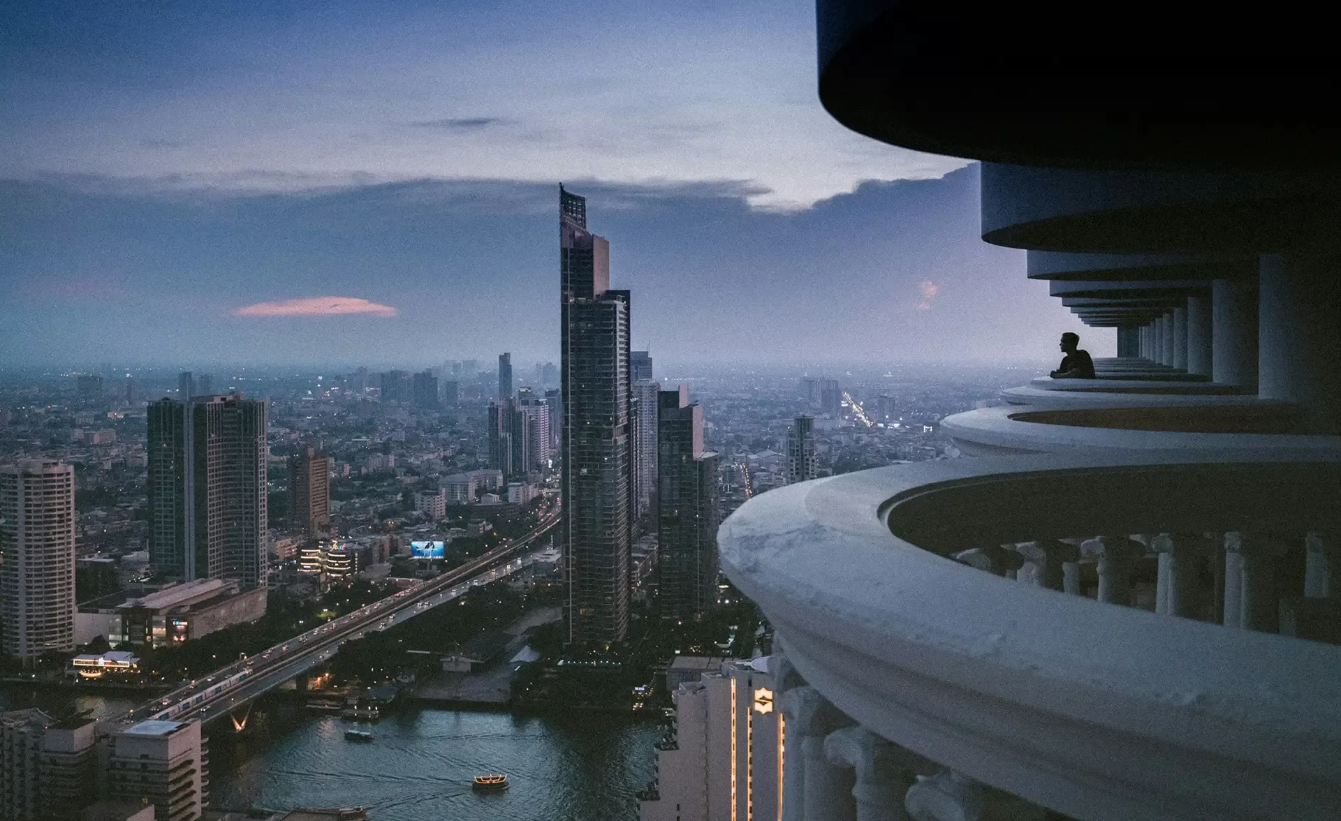 City skyline at dusk with a tall skyscraper and a river. A person sits on a balcony, overlooking the view. The scene captures the ambient glow of city lights and a hint of pink in the cloudy sky in Bangkok, Thailand.