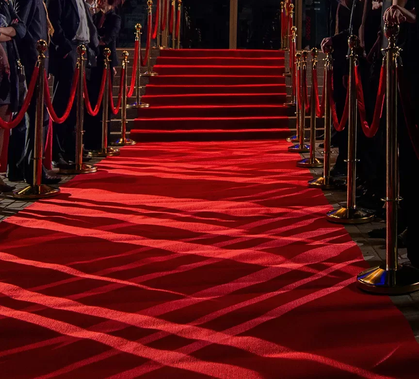 Red carpet event with a well-lit walkway and velvet ropes. People in formal attire line the sides, awaiting arrivals. Elegant dresses and suits are visible. Steps at the end of the carpet lead up, indicating a grand entrance.