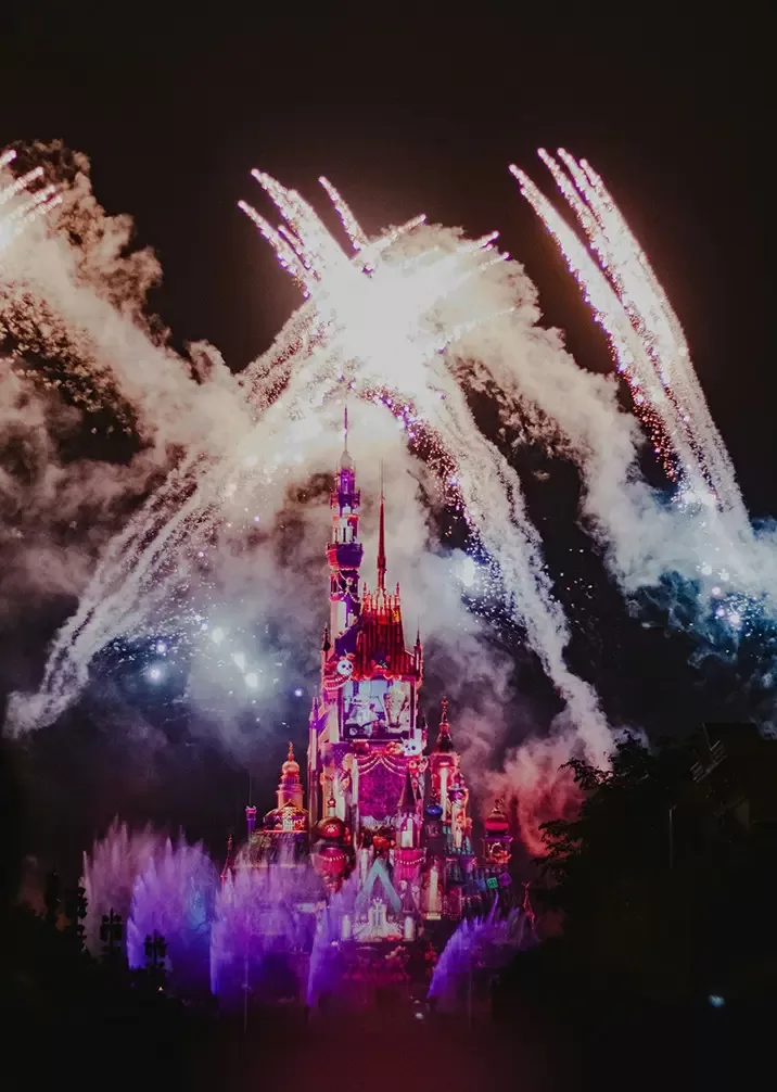 A colorful, illuminated castle at night with vibrant fireworks bursting in the sky above, creating a magical and festive atmosphere. The scene is lively, with bright lights and billowing smoke surrounding the castle.