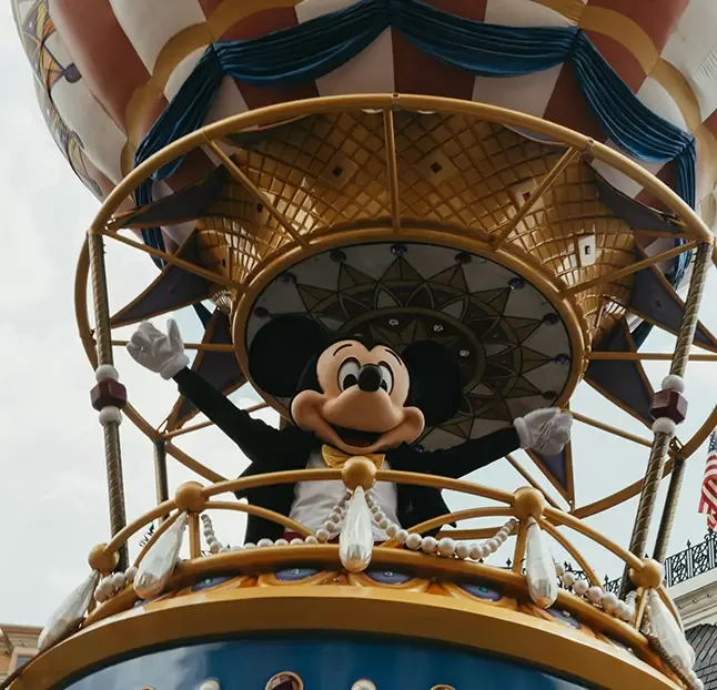 Character dressed as a famous cartoon mouse wearing a tuxedo, waving from a decorative hot air balloon basket with a colorful canopy. Background shows part of a building and sky.