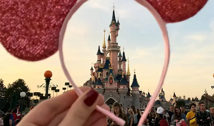 A hand holds pink sequined Mickey Mouse ears in front of a fairytale castle, with a large crowd of people in the background. The setting sun casts a warm glow over the scene, with trees and lampposts visible on the sides.
