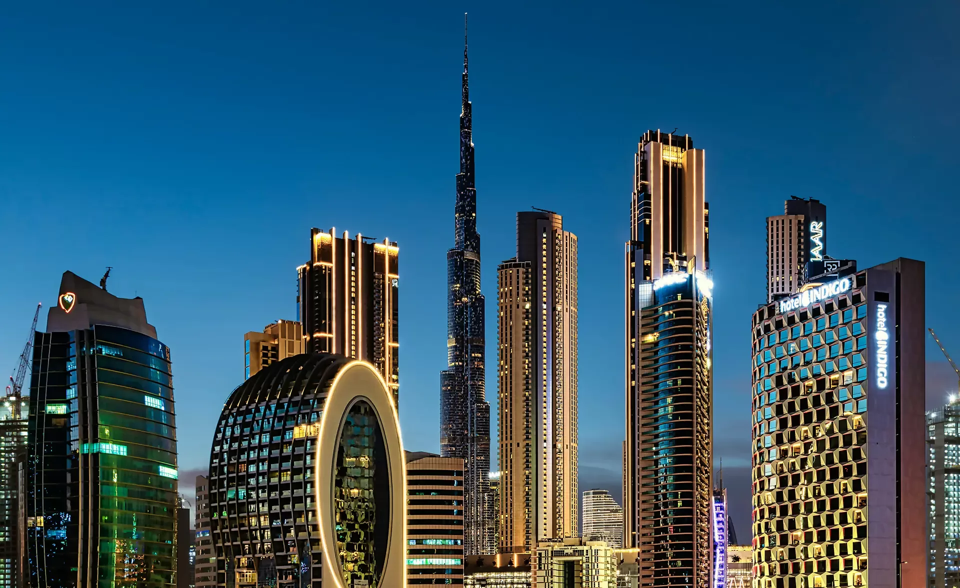 A nighttime view of a modern city skyline with tall, illuminated skyscrapers, including the Burj Khalifa and circular architectural designs.