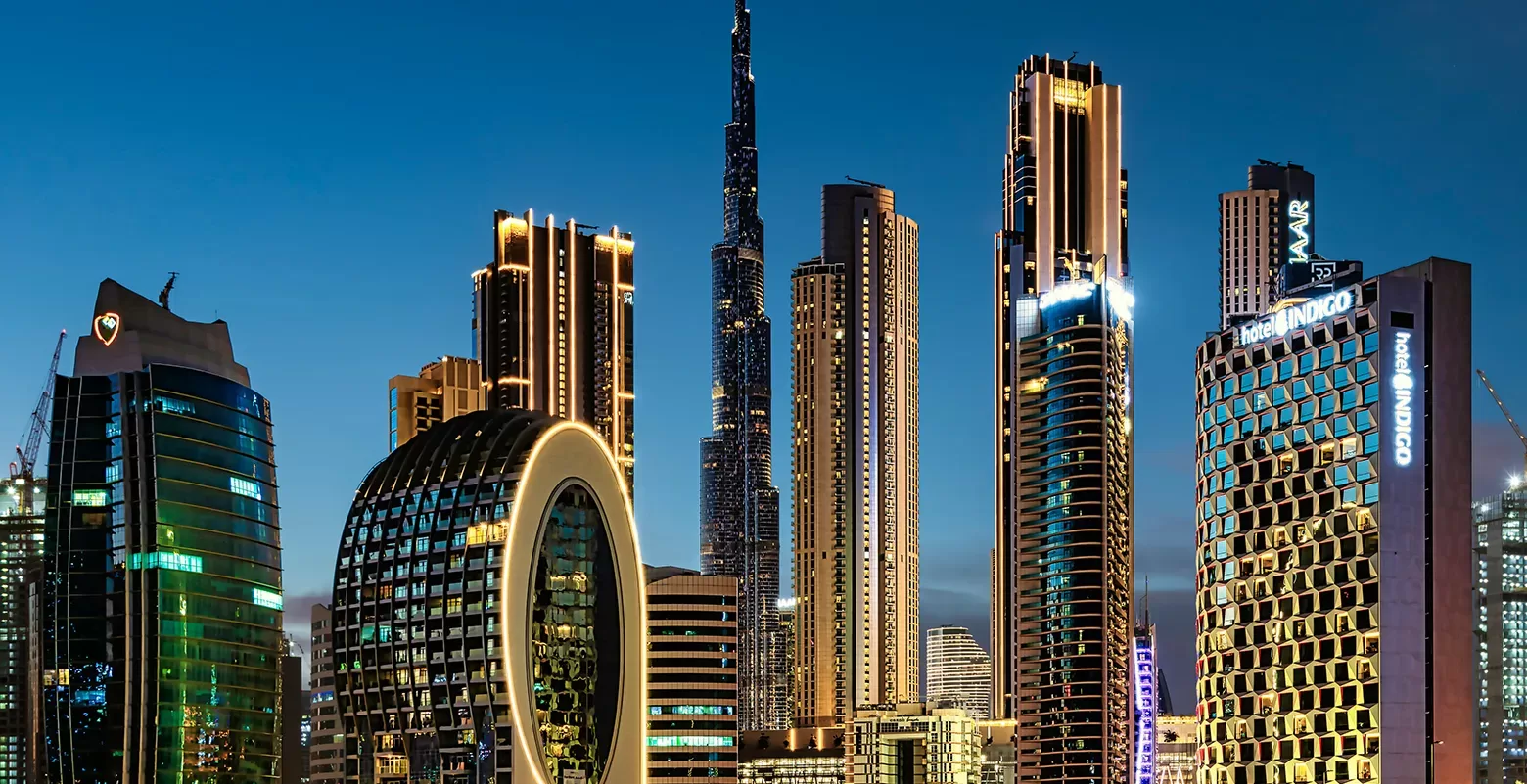 A nighttime view of a modern city skyline with tall, illuminated skyscrapers, including the Burj Khalifa and circular architectural designs.