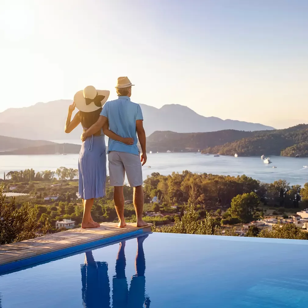 A couple stands arm in arm by an infinity pool, overlooking a scenic landscape with a lake, green hills, and distant mountains under a clear sky at sunset.