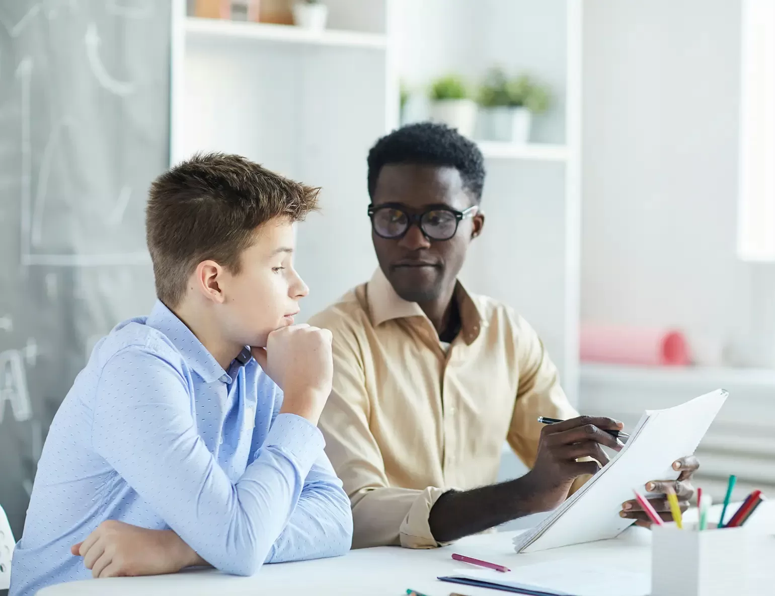 A young boy in a blue shirt sits thoughtfully at a desk, looking at a man beside him. The man, wearing glasses and a beige shirt, is holding a pen and notebook, appearing to discuss or explain something. Shelves and plants are in the background.