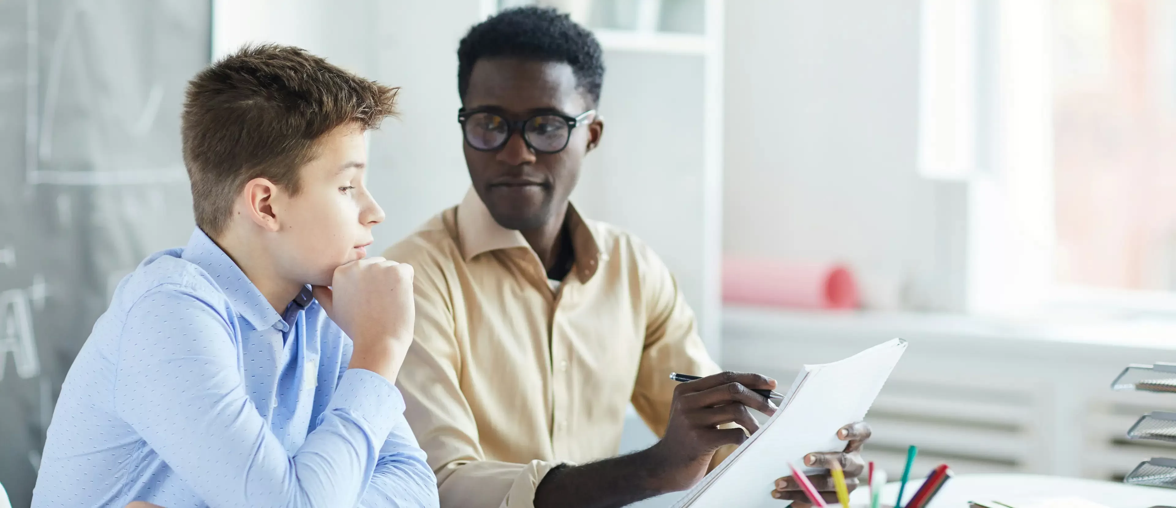 A teacher, holding a pen and paper, is sitting at a desk and talking to a student. The student is listening attentively with his hand on his chin. They are in a classroom with books and supplies visible.