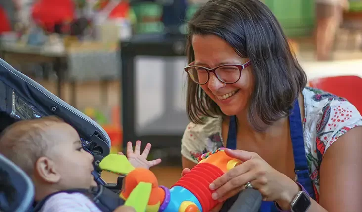 A woman with glasses smiles while interacting with a baby, who is seated. The baby reaches for a colorful toy held by the woman. The background is blurred, suggesting an indoor setting with vibrant decor by Quintessentially Foundation.