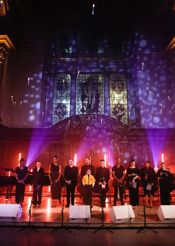 A choir stands in a dimly lit hall with ornate stained glass windows in the background. Colored lights cast patterns on the walls and ceiling, creating a vibrant atmosphere. A row of microphones is set in front of the singers at The Fayre of St James concert by Quintessentially Foundation.