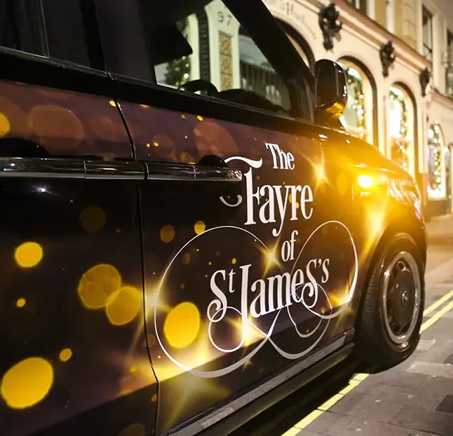 A black vehicle with festive golden lights displays the text The Fayre of St James on its side. The background shows a dimly lit street with decorated storefronts by Quintessentially Foundation.