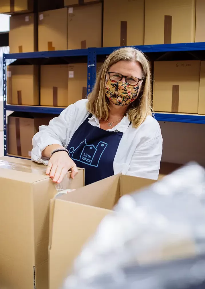 A woman wearing a colorful face mask and glasses stands in a storage room. She is packing cardboard boxes. Shelves filled with boxes are seen in the background by Quintessentially Foundation.